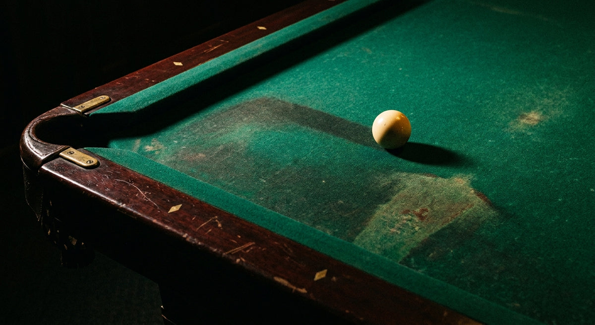 A close-up view of a worn pool table cloth surface under dramatic side lighting, highlighting texture and fading near a polished cue ball.